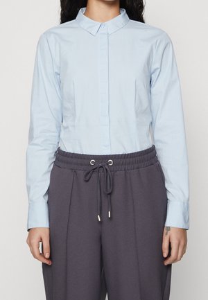 Žena oblečená v světle modré košili s knoflíky a límečkem, zastrčené do tmavě šedých kalhot se stahovací šňůrkou, stojící před neutrálním pozadím.