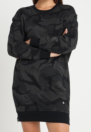 Donna che indossa un vestito felpa lungo con motivo mimetico nero e grigio scuro, con le braccia incrociate e polsini a coste.