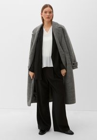 Long manteau en laine gris sur un blazer noir et un haut plissé blanc, associé à un pantalon large noir et des chaussures noires brillantes. Fond uni.