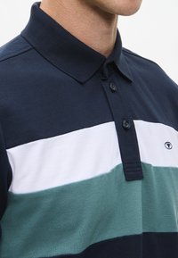 Tummansininen polo-paita, jossa on vaalean ja teal-värin vaakaraitoja, kaulus ja napitusyksityiskohdat.