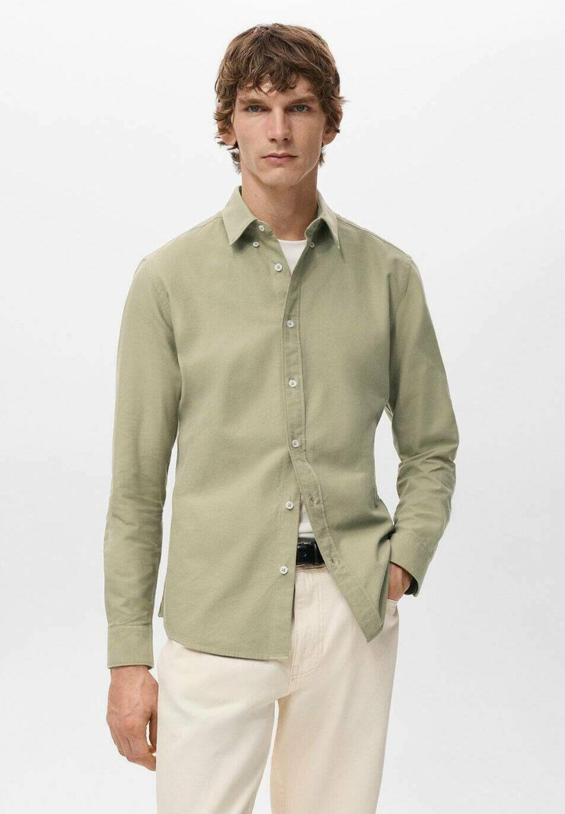 Camisa de manga larga color verde claro con botones y cuello. Hecha de tela con textura suave y brillo sutil. Combinada con pantalones de color crema.