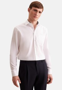 Hvid langærmet skjorte med knapper og klassisk krave, enkelt brystlomme og subtile mønstrede detaljer på den indvendige krave, parret med mørke bukser.