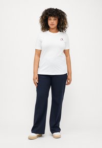 Λευκό βαμβακερό T-shirt με κοντά μανίκια, που διαθέτει ένα μικρό μαρoon λογότυπο στο αριστερό στήθος, σε συνδυασμό με ναυτικές παντελόνες σε φαρδιά γραμμή και μπεζ υποδήματα.