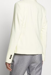 Jasnożółta dopasowana marynarka o gładkiej fakturze, z pojedynczym wentylem i detalem trzech guzików na rękawach; zestawiona z teksturowanymi srebrnymi spodniami.