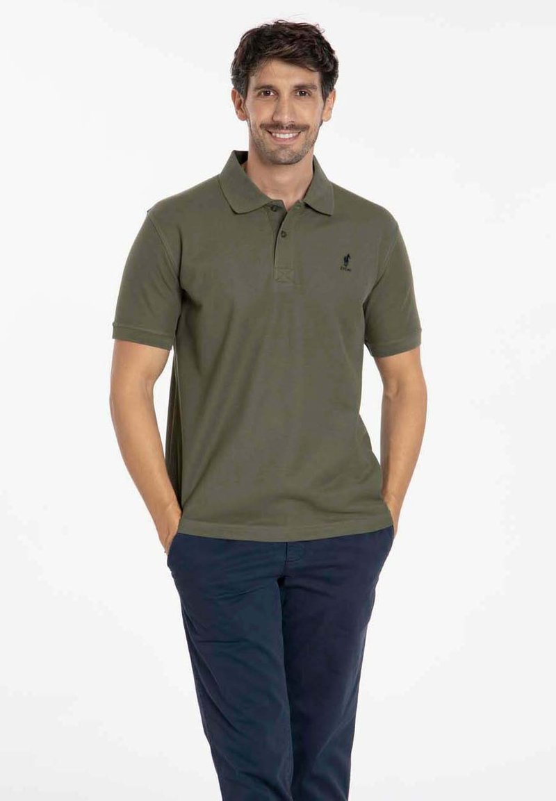 Polo shirt vert olive avec col, manches courtes et un petit logo brodé sur la poitrine, associé à un pantalon bleu marine foncé.