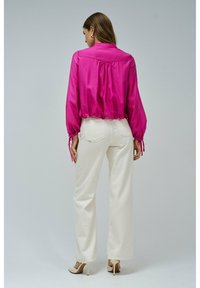 Donna che indossa una blusa lunga a maniche con un vivace colore rosa, pantaloni bianchi a vita alta e larghi, e tacchi alti beige, in piedi di spalle.