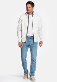 Witte ritsjack met zwarte strepen, gedragen over een wit shirt en lichtblauwe jeans. Beige instap schoenen maken de outfit compleet. Casual ontwerp.