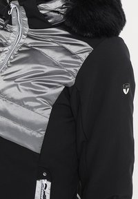 Schwarz-silberne isolierte Jacke mit glänzender, gesteppten Oberfläche, ausgestattet mit einem kapuzen mit Pelzfutter und Reißverschlusstaschen. Akzente setzen ein Logo-Patch.