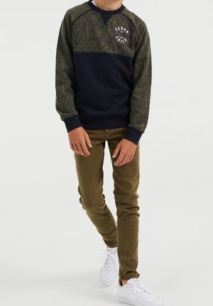 Sudadera azul marino y verde oliva con mangas estampadas de hojas, cuello redondo y gráfico frontal; combinada con pantalones beige ajustados y zapatillas blancas.