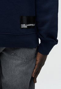 Κλείσιμο του προσώπου ενός ατόμου που φοράει μπλε μπλούζα με κουκούλα με μαύρη ετικέτα «KARL LAGERFELD JEANS» και ξεβαμμένο γκρι τζιν που δείχνει την πίσω τσέπη.