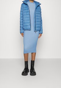 Jasnoniebieska pikowana kurtka z kapturem, noszona na dopasowanej prążkowanej sukience midi. Stylizację dopełniają czarne masywne botki za kostkę.