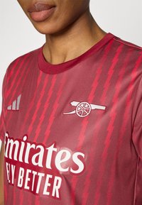 Μπορντό αθλητική φανέλα με κόκκινες διαγώνιες ρίγες, με λευκό κεντημένο λογότυπο συλ�λόγου και τη φράση "Emirates Fly Better" στο μπροστινό μέρος.