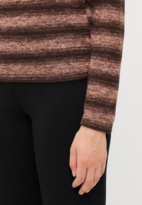 Ruskea ja beige raidallinen neulepaita pitkillä hihoilla. Sileä rakenne, tiukasti istuva malli, puettuna mustien housujen kanssa.
