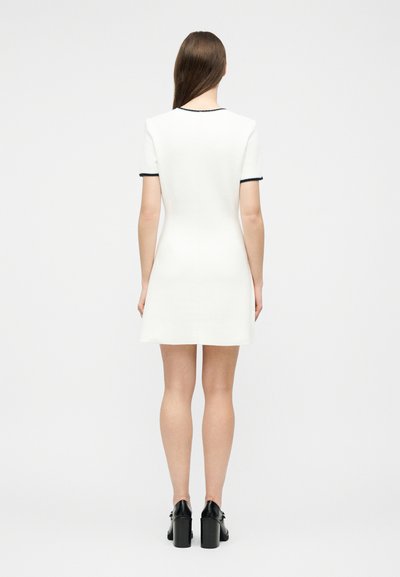 Γυναίκα με μακριά καστανά μαλλιά, που φορεί κοντό λευκό φόρεμα με μαύρο περίγραμμα και μαύρα τακούνια, στέκεται με την πλάτη προς τα εμπρός σε ένα απλό φόντο.