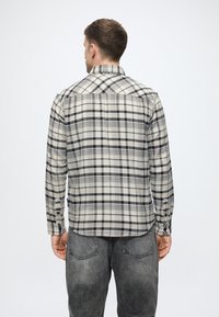 Beżowo-czarna koszula w kratę z długim rękawem, z klasycznym kołnierzykiem, mankietami na guziki i zaokrąglonym dołem. Noszona z ciemnymi dżinsami.