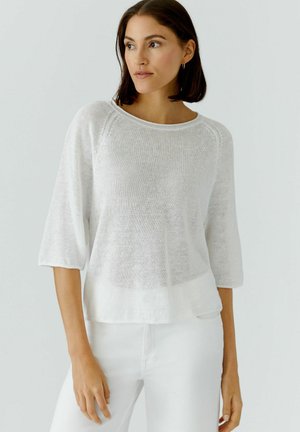 Vrouw met donker schouderlang haar, gekleed in een witte losgebreide trui met driekwart mouwen en een witte broek, kijkt opzij.