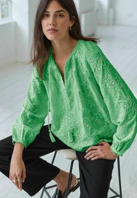 Blusa verde ricamata con scollatura a V, motivi floreali, maniche a sbuffo e decorazioni con lacci, abbinata a pantaloni neri.