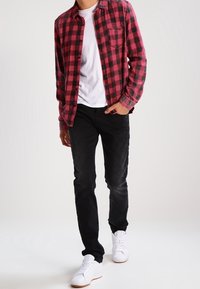 Rood en zwart geruite button-up shirt over een wit t-shirt, gecombineerd met zwarte jeans en witte sneakers. De stof lijkt zacht en heeft een casual pasvorm.