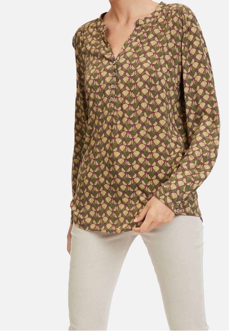 Femme portant un chemisier à manches longues à motifs avec des formes géométriques vertes, beiges et violettes et un pantalon beige clair, main reposant près de la hanche.