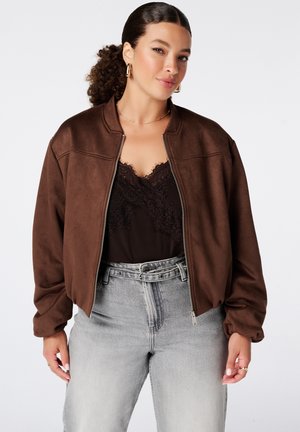 Femme aux cheveux bouclés attachés porte une veste marron zippée, un top en dentelle noire, un jean gris clair taille haute, des boucles d'oreilles créoles dorées et un collier.