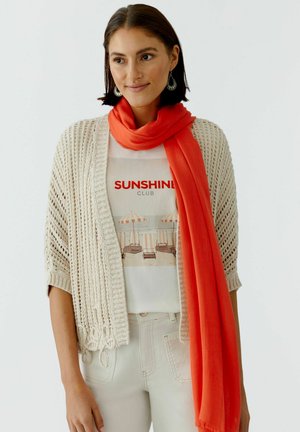 Frau trägt eine beige Strickjacke, ein weißes Grafik-T-Shirt mit dem Aufdruck "Sunshine Club", beige Hosen, einen orangen Schal und hängende Ohrringe, lächelt sanft.