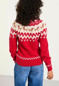 Frau mit lockigem Haar, die einen roten Pullover mit Rentieren und geometrischen Mustern trägt, kombiniert mit blauen Jeans, und von der Kamera abgewandt ist.