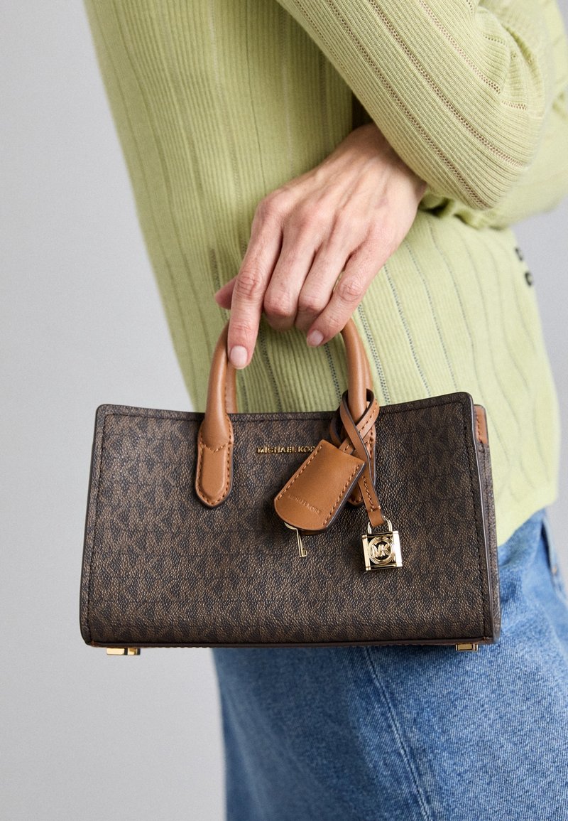 Main tenant un petit sac à main marron Michael Kors avec des anses en cuir et un charm cadenas doré, portant un pull côtelé vert et un jean bleu.