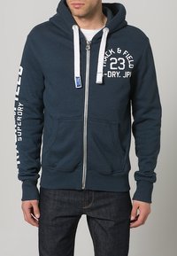 Hombre que lleva una sudadera con cremallera de color azul oscuro con cordones blancos y el texto "Track & Field 23" en el pecho y "Superdry" en la manga.