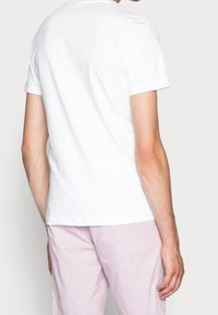 Bílý bavlněný tričko s kulatým výstřihem a krátkými rukávy, přiléhavý střih, hladká textura, z pohledu zezadu. Nosené s světle růžovými šortkami.