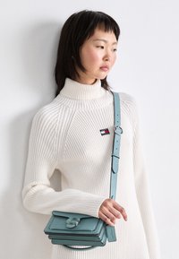 Suéter blanco de cuello alto tejido con textura acanalada. Pequeño bolso de cuero en color teal cruzado con compartimentos apilados y un cierre circular.