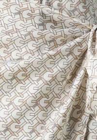 Krém színű textil, amelyen ismétlődő geometrikus minta látható világosbarnában, a "GUESS" szó beágyazva a dizájnba. Simás tapintású.