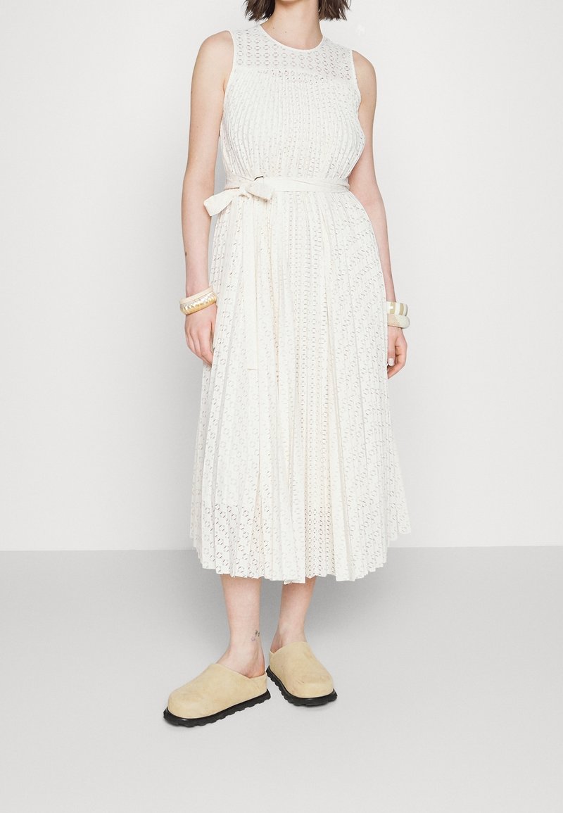 Robe blanche sans manches en tissu texturé avec des découpes circulaires, dotée d'un corsage ajusté et d'une ceinture à la taille, assortie à des sabot beiges.