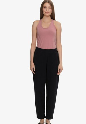 Młoda kobieta stoi, ubrana w różowy, bezrękawnikowy top i luźne czarne spodnie na jednolitym białym tle.