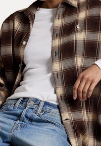 Brun och beige rutigt knapp-up skjorta i lager över en vit t-shirt, tillsammans med ljusblå denimjeans. Texturerad tyg och synliga knappar.