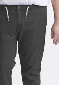Mørkegrå chinos med en glatt tekstur, knappestenging og hvite snor med svarte detaljer i midjen.
