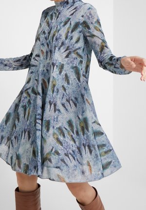 Vzdušné dlouhé šaty s modrým, zeleným a hnědým abstraktním vzorem, nošené s hnědými botami, ruce mírně rozpažené, jako při otáčení.