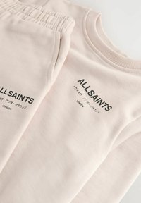 Hellbeige Sweatshirt und passende Hose mit elastischem Bund, die den Schriftzug "ALLSAINTS" in Schwarz trägt. Glatte, weiche Stoffoberfläche.
