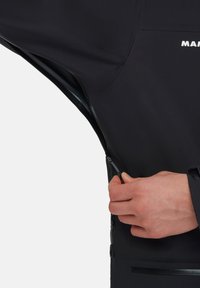 Schwarze wasserdichte Jacke mit einem Reißverschluss, glatter Textur, elastischem Material und einem Logo auf der Oberseite. Hand in der Nähe des Reißverschlusses platziert.