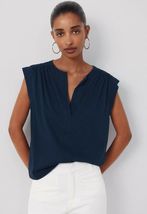 Vrouw met donker haar in een marineblauwe mouwloze top en witte broek staat tegen een effen lichte achtergrond, met grote oorbellen in de vorm van hoepels.