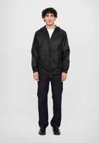 Veste légère noire avec fermeture éclair à l'avant, poignets élastiques et capuche. Associée à un jean en denim foncé et des chaussures noires.