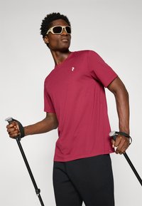 Maroon short-sleeve t-shirt with a crew neckline and a small white logo on the chest. Worn with black pants and holding a trekking pole.
