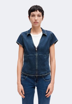Un mannequin aux cheveux courts et foncés porte un haut en denim à manches courtes, zippé sur le devant, par-dessus une chemise blanche, avec un jean assorti, sur un fond clair uni.