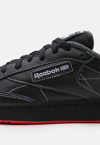 Černé kožené tenisky s bílým prošíváním, texturovanou boční částí, logem Reebok a červenou gumovou podešví. Mají šněrovací design a šedé akcenty.
