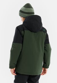 Grüne und schwarze wasserdichte Jacke mit Kapuze, elastischen Bündchen und einem Logo auf der Schulter, kombiniert mit einer grün gestreiften Strickmütze.