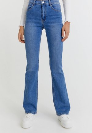 Person wearing high-waisted blue bootcut jeans, white long-sleeve top, and white sneakers, standing against a light background.