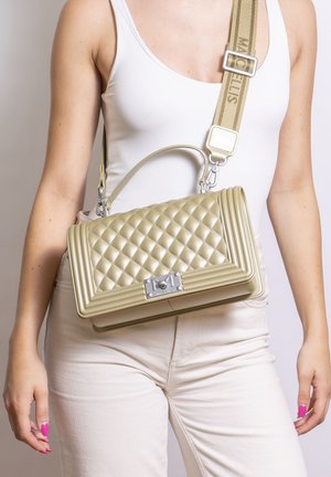 Femme portant un débardeur blanc et un pantalon avec un sac à main beige matelassé, doté d'une large bandoulière portée à l'épaule et d'une fermeture argentée.