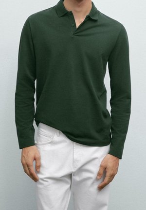 Homme portant un polo à manches longues vert foncé et un pantalon blanc, debout devant un fond uni gris clair.