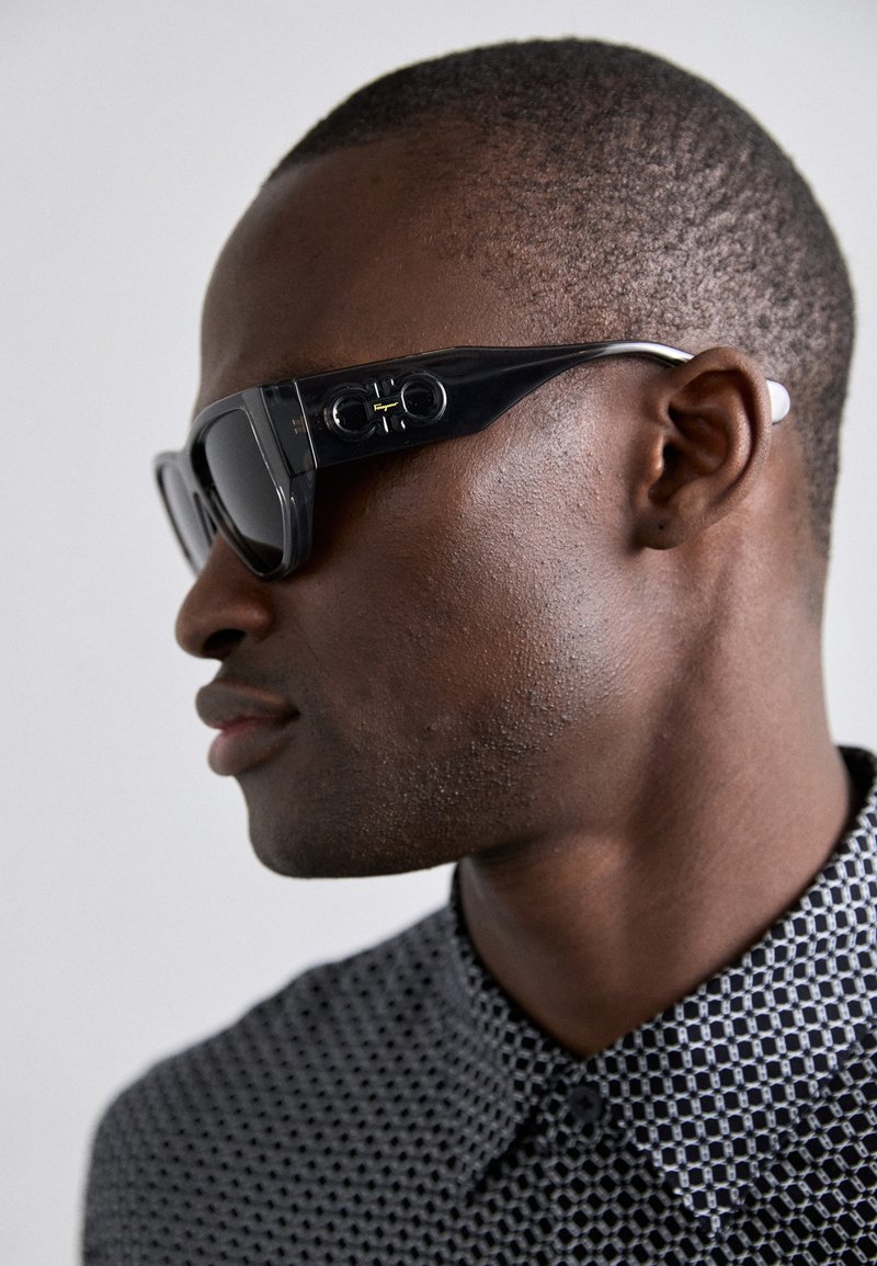 Fekete műanyagból készült napszemüveg, amelynek letisztult dizájnja van. Sötét lencsékkel, ezüst szárakkal és kör alakú díszítésekkel rendelkezik a keretén.