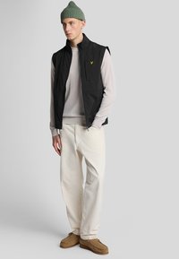 Schwarze ärmellose Weste mit gelbem Logo, über einem hellgrauen Pullover getragen, kombiniert mit beigen Hosen und braunen Schuhen. Grüner Strickmütze.
