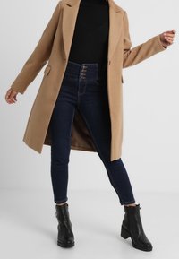 Kamelfrakke med skræddersyet design, sort ribbet sweater, højtaljede mørke denim skinny jeans og sorte ankelstøvler med en chunky hæl.